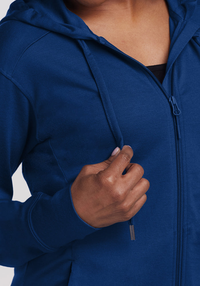 A person wearing the Woolx Ryann Hoodie in Estate Blue holds a drawstring with their right hand. Only their torso, hand, and part of their face are visible. This lightweight hoodie is perfect as a travel essential.