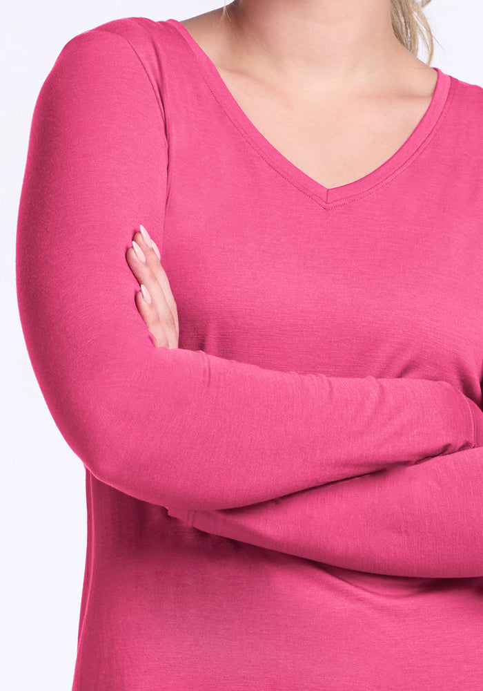 A person stands with arms crossed wearing the Woolx Millie Long Sleeve in Raspberry Fizz, a bright pink, moisture-wicking Merino wool top with long sleeves and a V-neck. The cropped image shows their torso and part of their face against a plain light background.