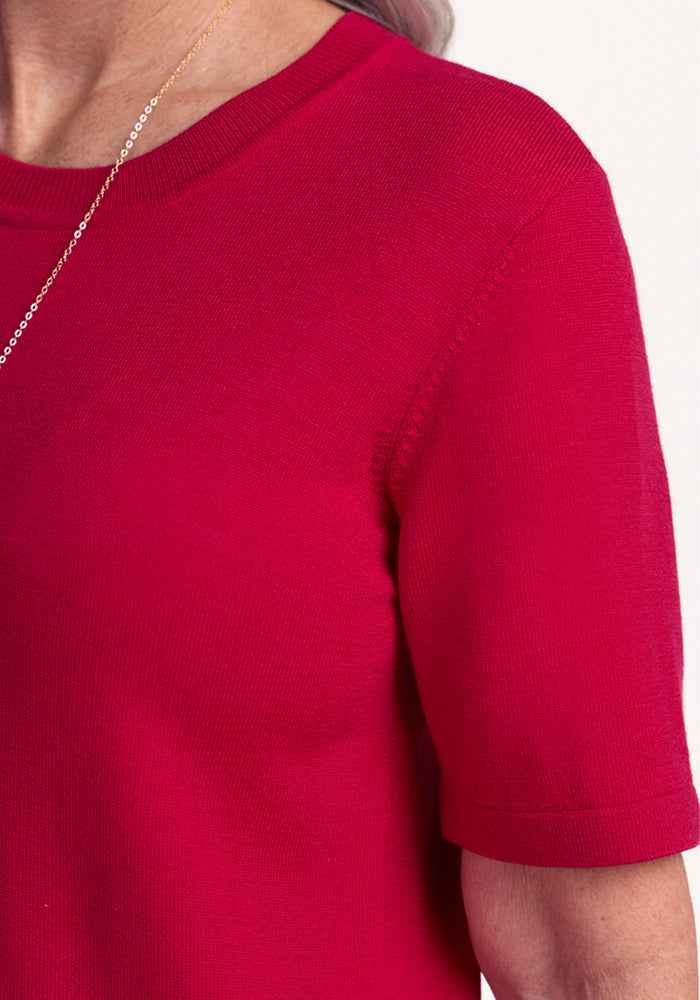 A close-up of a person wearing the Woolx Anna Crew Sweater in Poppy Spark and a delicate gold chain necklace, with part of their neck, shoulder, and arm visible against a white background.