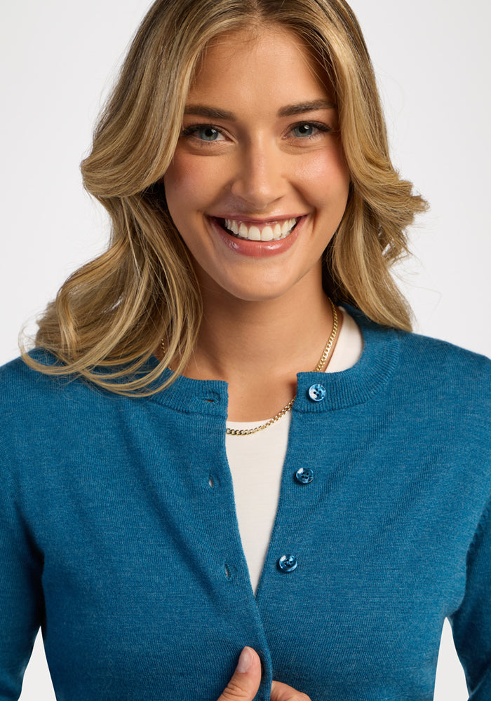 Smiling woman in Woolx Caitlin Sweater - Blue Pine over a white shirt, wavy blond hair, gold chain necklace.