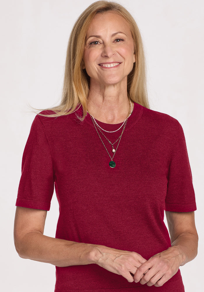 Smiling woman with long blonde hair in a Woolx Anna Crew Sweater - Beet Red, layered necklaces, white background.
