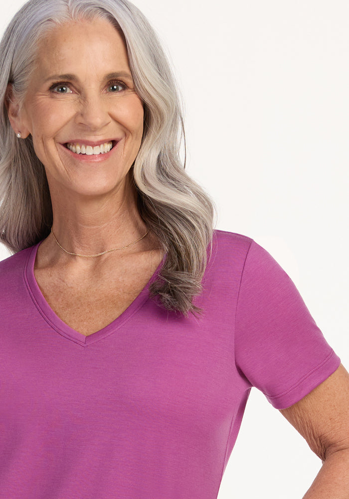 Smiling older woman with long gray hair wears the Woolx Junie V Neck in Purple Wine, a relaxed fit Merino wool tee, standing against a plain light background.