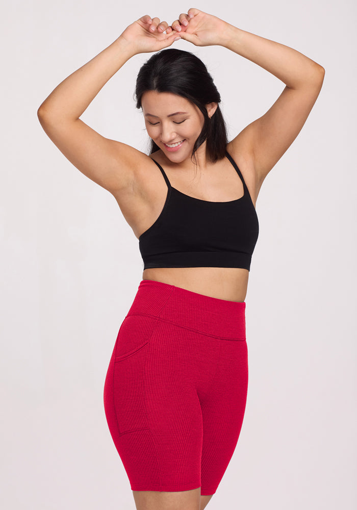 A woman with dark hair smiles and raises her arms, wearing a black crop top and Woolx Miley Bike Shorts - Poppy Spark. The high-waisted red ribbed shorts with pockets stand out against a plain light background.