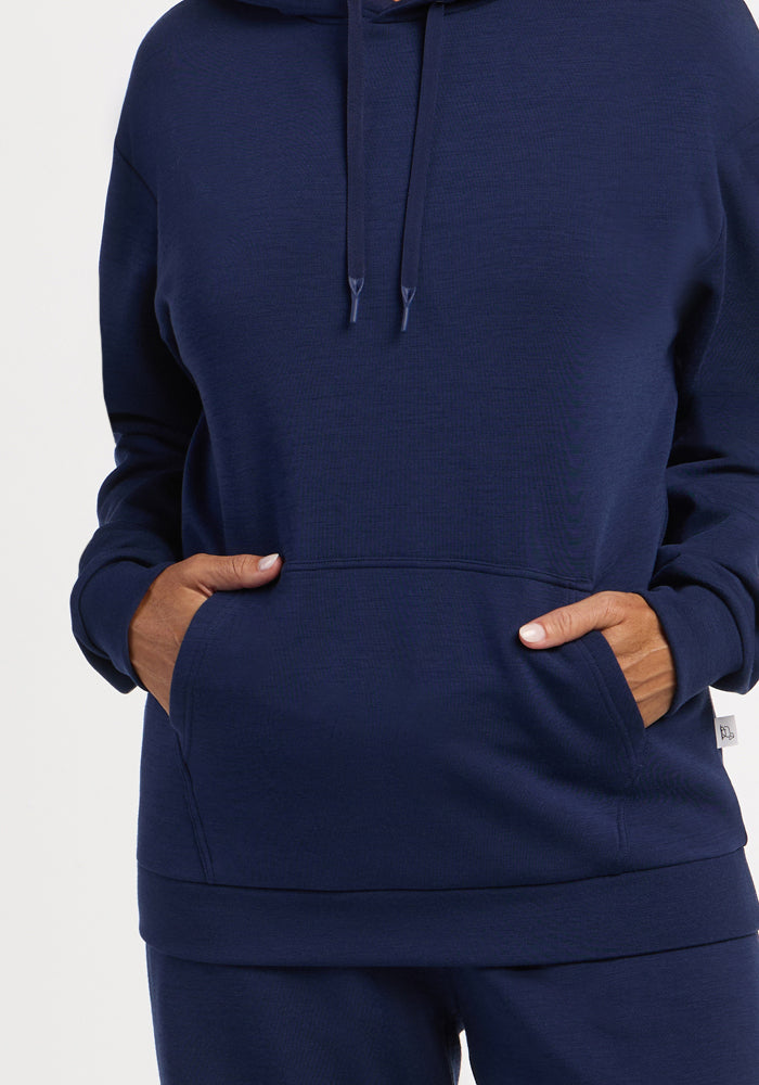 Model in a Woolx Avery Hoodie - Ocean Cavern, hands in pocket, stands before a plain light background.