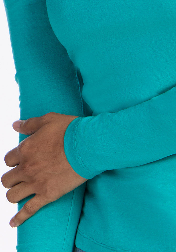 A person wearing the Woolx Remi Long Sleeve T-Shirt in Cool Jade crosses one arm over their torso and gently holds their opposite arm, set against a plain white background.
