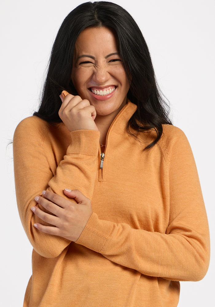 Woman beams in the Woolx Collins Half Zip Sweater in Butterscotch, holding its collar with arms crossed.
