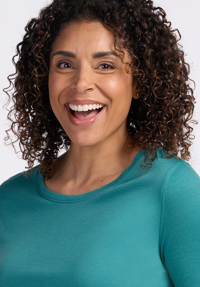 A woman with curly hair smiles at the camera, wearing the Woolx Jenny 3/4 Sleeve Crew Neck - Surfside, against a plain light background.