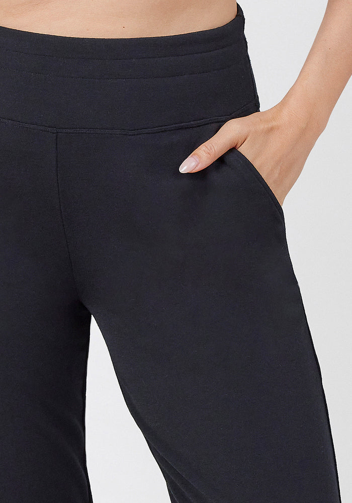 Wearing Woolx Madden Joggers - Black with a wide waistband, a person places their right hand in the pocket, revealing part of their bare midriff and arm.