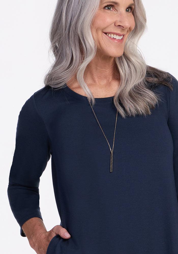 Smiling older woman with long gray hair wears the Woolx Rissa A Line Dress in Deep Navy and a long pendant necklace, posing with one hand in her pocket—perfect for fall and winter layering against a light background.