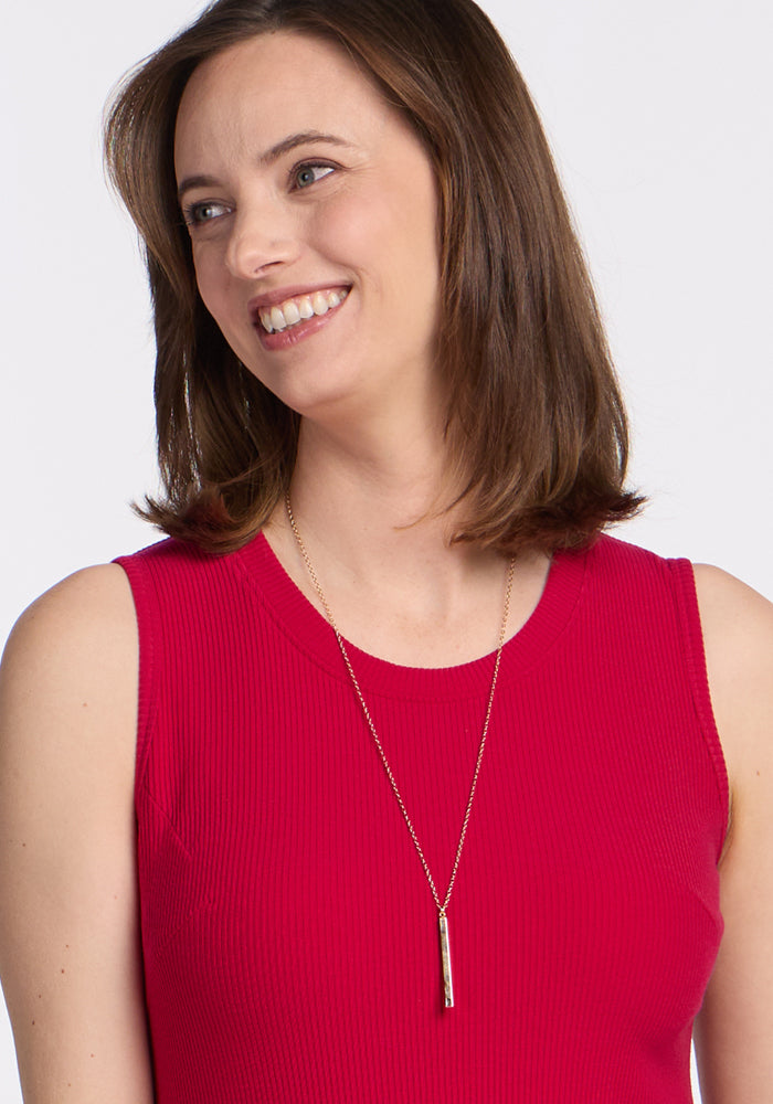 A woman with brown hair is smiling and looking to the side against a white background while wearing the Woolx Iris Ribbed Dress in Poppy Spark and a long gold necklace.