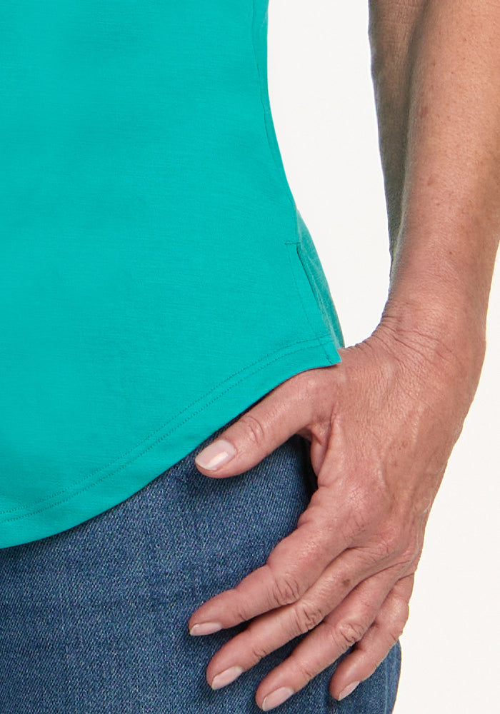 A close-up of a person's hand resting on their hip, wearing the Woolx Bella Tunic Top in Cool Jade and blue jeans, with a plain light background.