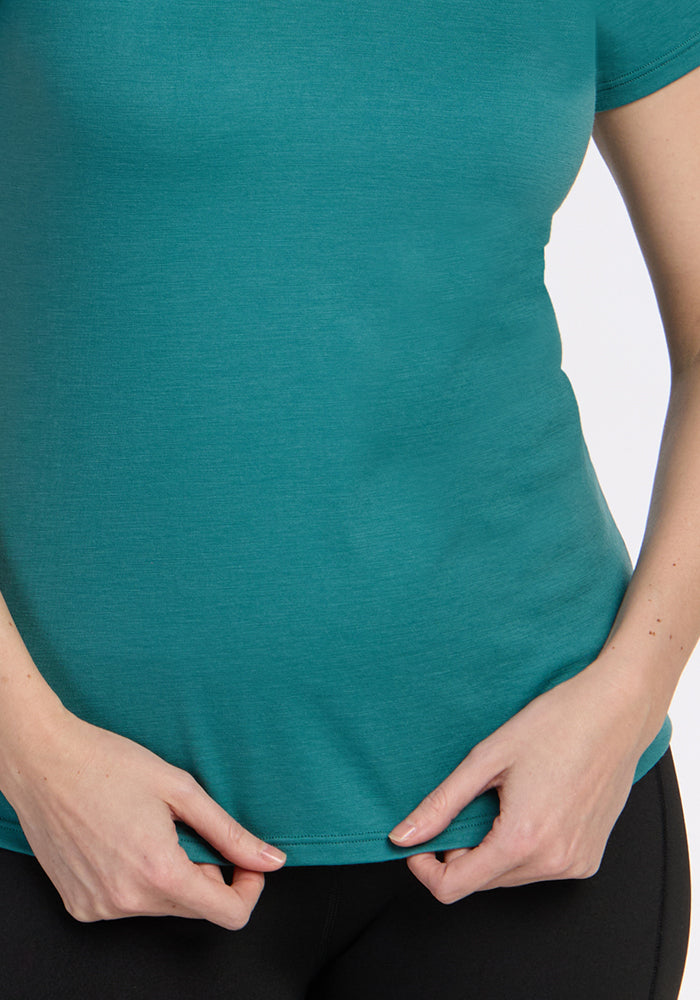 A person wearing the Woolx Mia V Neck - Surfside, a fitted teal Merino wool t-shirt, and black pants holds the bottom edge of their shirt with both hands against a plain background.