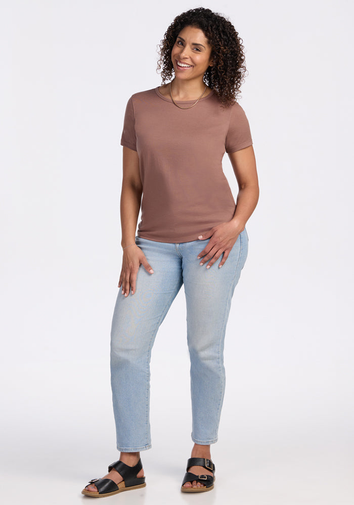 A woman with curly hair smiles with one hand on her hip, wearing the Woolx Liza Crew Neck Tee in Sienna Kiss, light blue jeans, and black sandals against a plain white background.