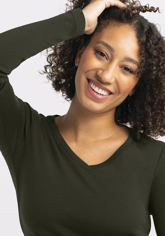 Smiling woman wears the Woolx Eva Tunic - Duffel Bag, hand to head, showing its dark green V-neck style.