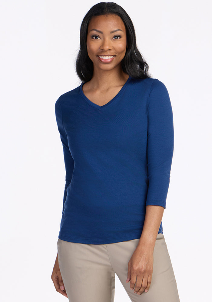 A woman with long black hair, dressed in a Woolx Elena 3/4 Sleeve V Neck top in Estate Blue and beige pants, smiles while standing against a plain white background. 