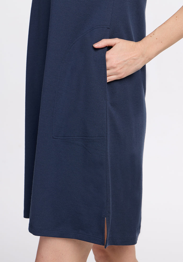 A person wearing the Woolx Georgie Dress in Deep Navy, featuring wrinkle-resistant fabric, a large side pocket, and a small slit near the hemline, rests their hand inside the pocket.