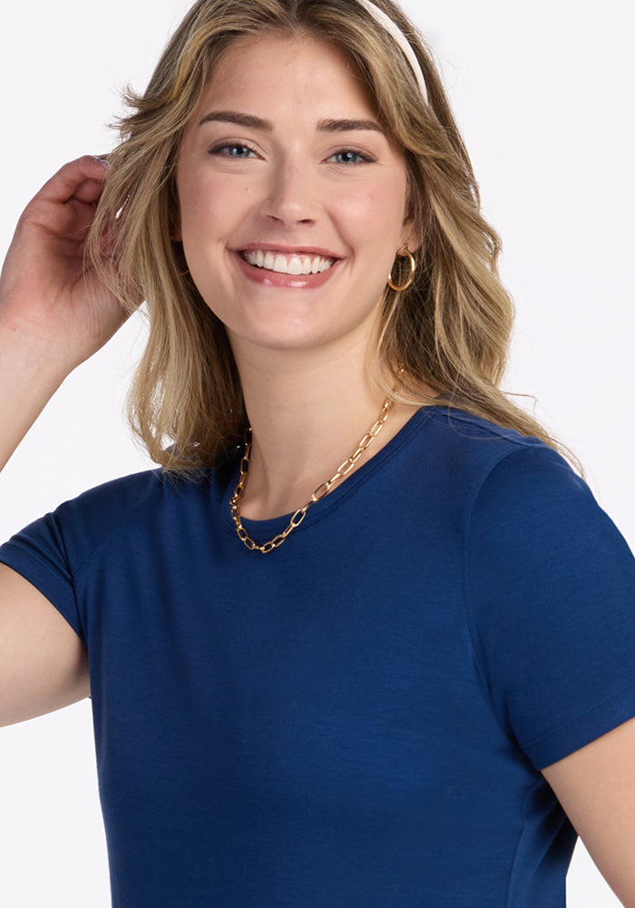 A smiling blonde woman wears the Woolx Liza Crew Neck Tee in Estate Blue, paired with gold hoop earrings and a gold chain necklace. She raises one hand near her head against a plain white background.