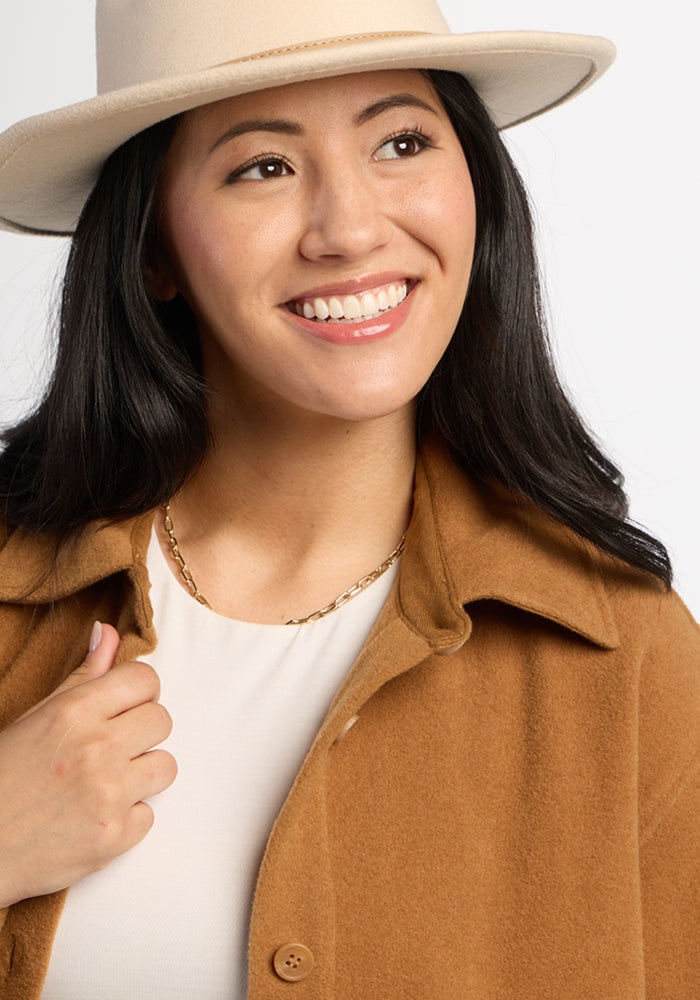 Woman smiling in Woolx Jovie Shacket - Saddleback, beige hat, brown coat & gold necklace against a light background.