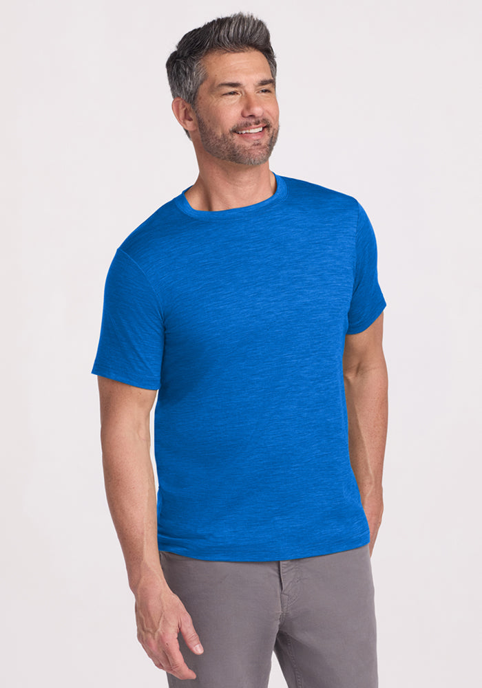 A man with short gray hair and a beard smiles while wearing the Woolx Endurance Tee in Glacial Blue and gray pants, standing against a plain, light-colored background.