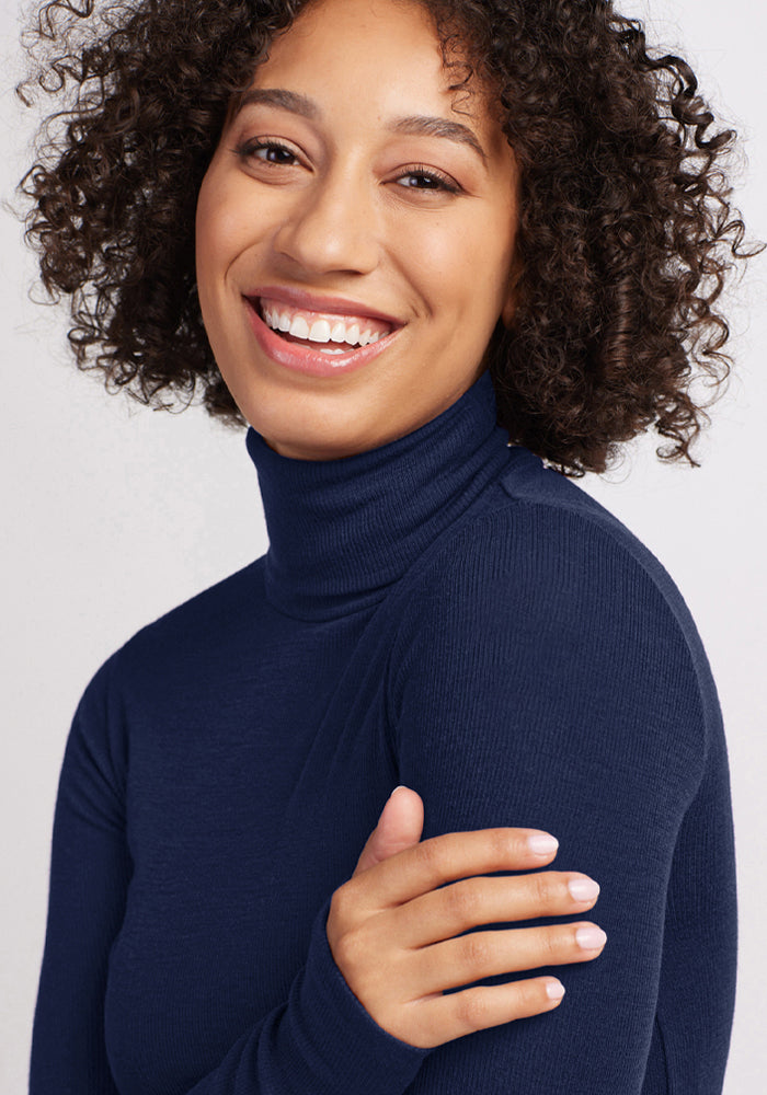 Curly-haired woman smiles in a Woolx Sage Ribbed Turtleneck - Ocean Cavern, her right hand on her left arm.