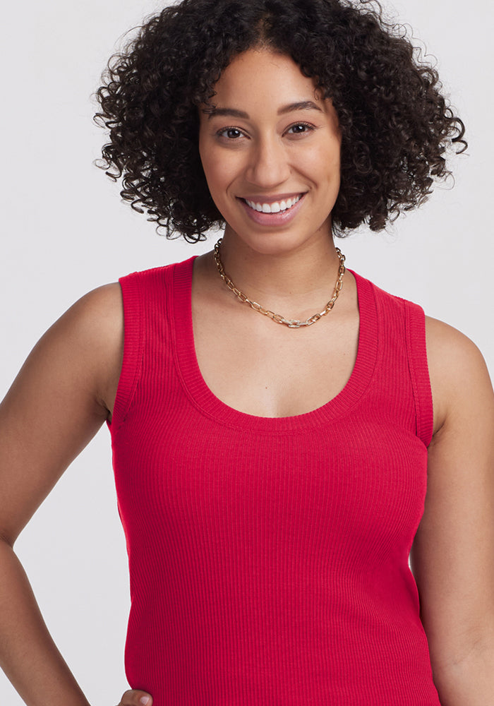 A woman with curly dark hair smiles at the camera, wearing the Woolx Sloane Ribbed Tank in Poppy Spark and a gold chain necklace against a plain light background.