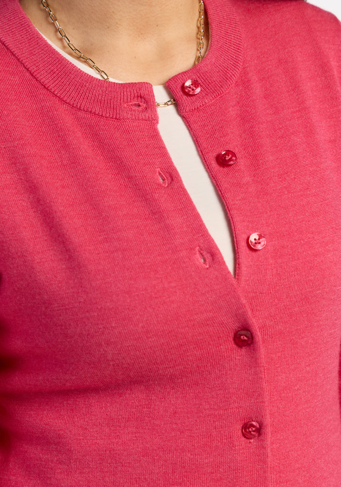 A woman wears the Woolx Caitlin Sweater in Vivid Rose, unbuttoned at the top, over a white shirt and gold chain.
