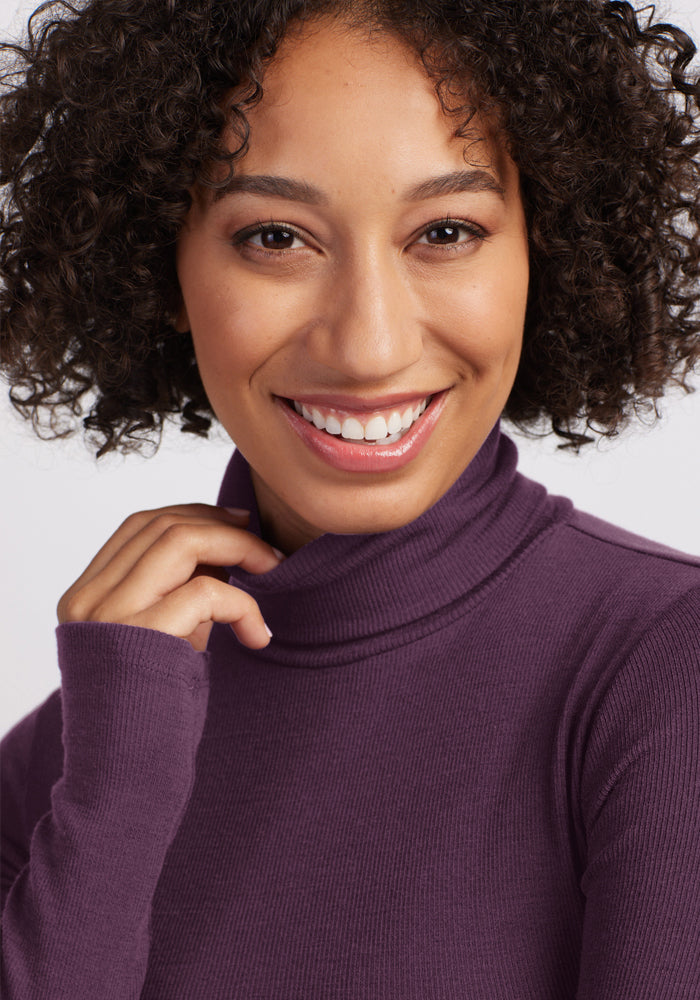 Smiling person in Woolx Sage Ribbed Turtleneck - Purple Iris, hand on collar, curly hair, white background.
