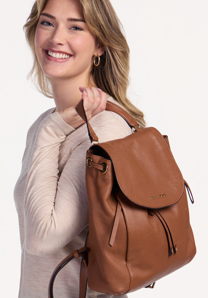 A smiling woman with wavy blonde hair wears the Woolx Millie Long Sleeve in Cream Heather and holds a brown leather backpack over her shoulder, looking at the camera against a plain light background.
