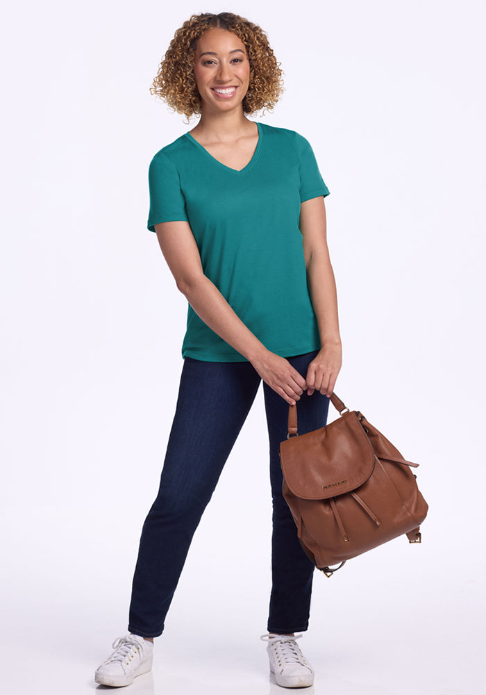 A smiling woman with curly hair wears the Woolx Junie V Neck - Surfside tee, dark jeans, and white sneakers. She stands holding a brown leather backpack in front of her against a plain white background.