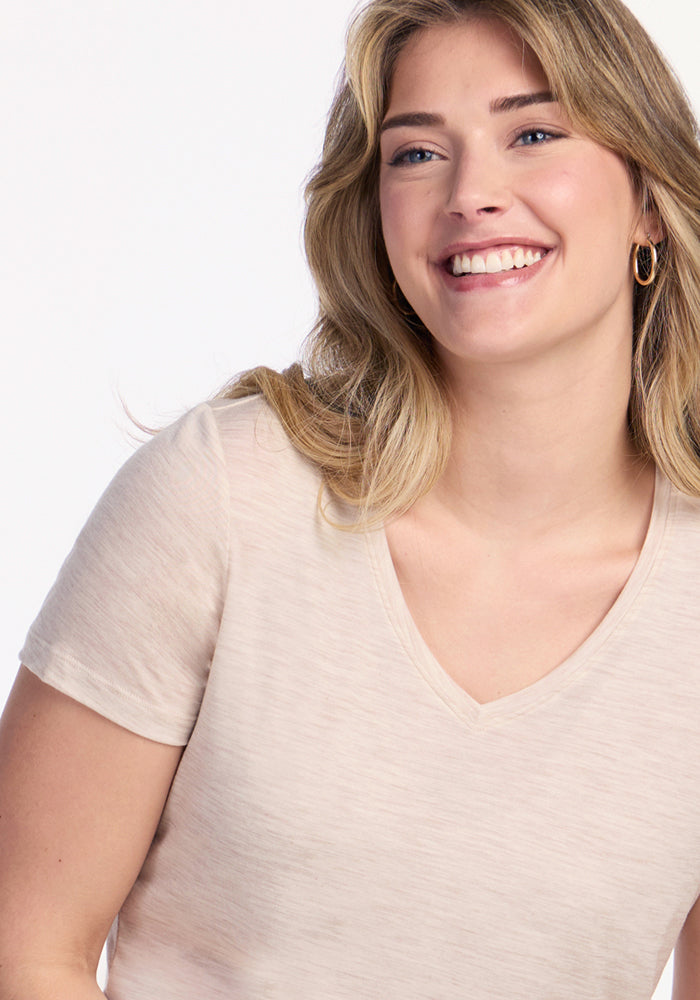 A smiling woman with wavy blonde hair wears the Woolx Junie V Neck in Cream Heather and gold hoop earrings, posing against a plain white background.