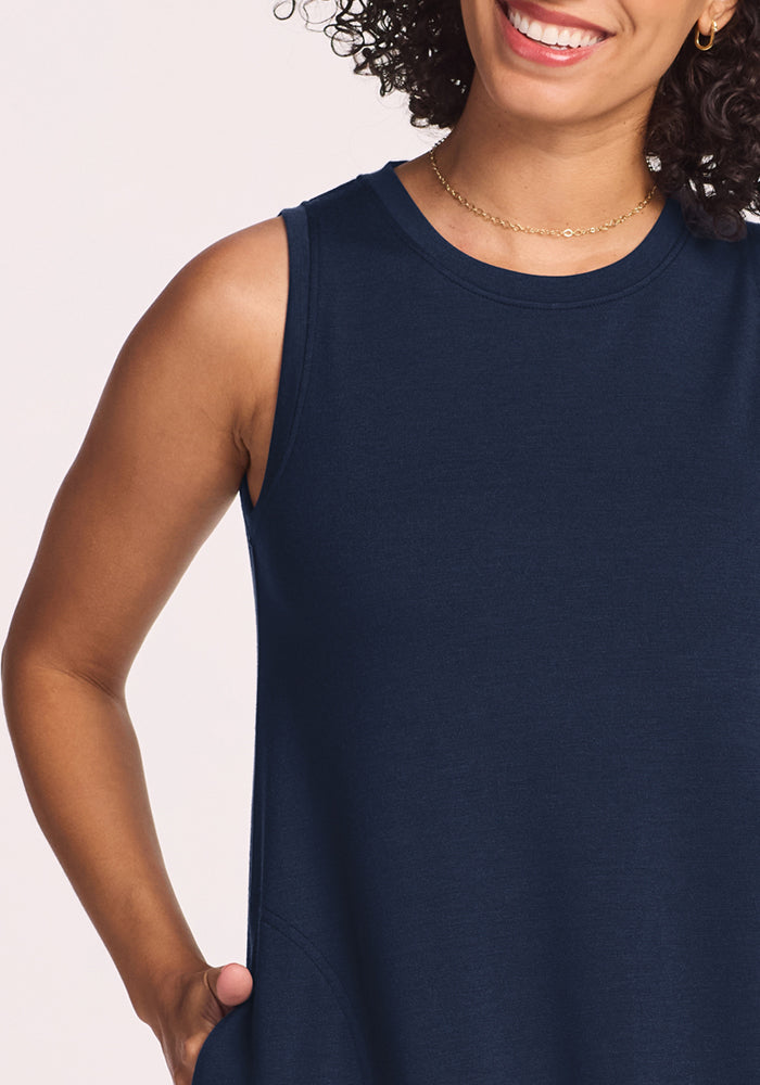 A woman with curly hair and a gold necklace smiles, showing her teeth, with one hand in the pocket of her Woolx Clara Dress in Deep Navy. The sleeveless Merino wool dress stands out against a light background.