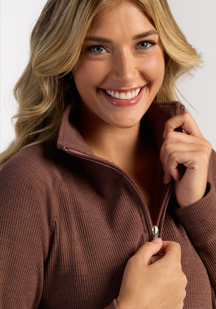 A smiling woman with wavy blond hair zips up the Woolx Quinn Ribbed 1/4 Zip in Sable Heather, facing the camera.