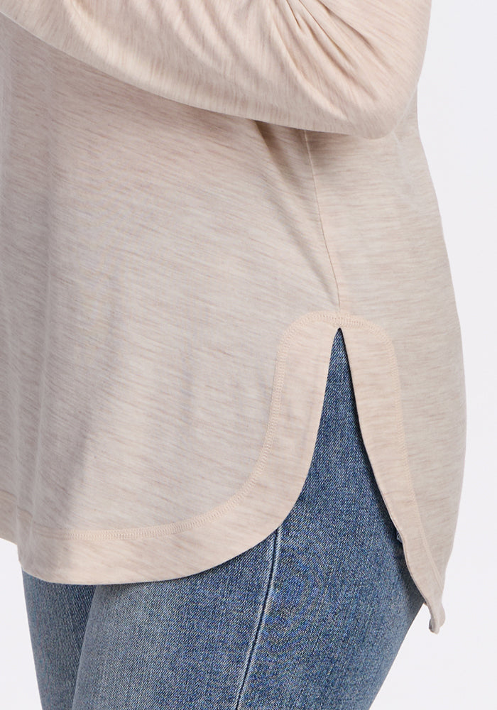 A close-up of a person wearing light blue jeans and the Woolx Hazel Tunic in Cream Heather, a lightweight merino wool top featuring a rounded hem and side slit.