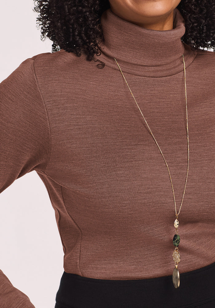 Woman wears the Woolx Peyton Turtleneck in Sable Heather with a gold necklace and black pants for a chic, cozy vibe.