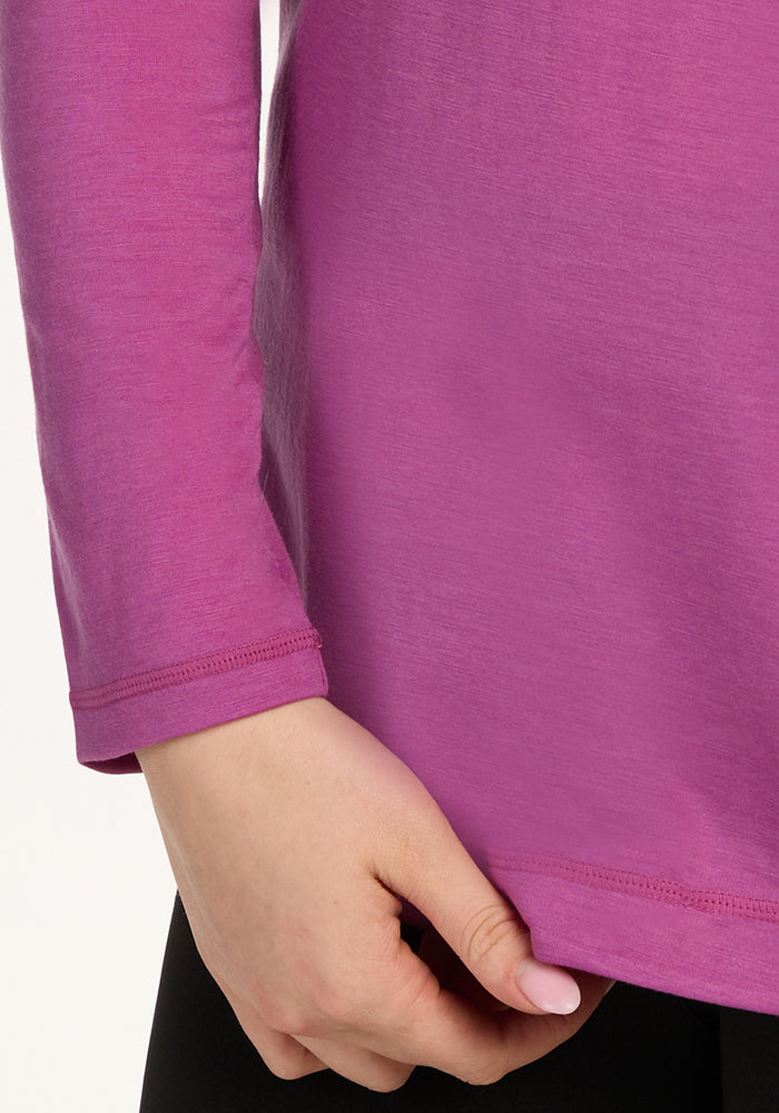 A close-up of a person wearing the Woolx Hazel Tunic in Purple Wine with black pants. Their left hand holds the hem of the long-sleeved merino wool top. The background is plain and light-colored.
