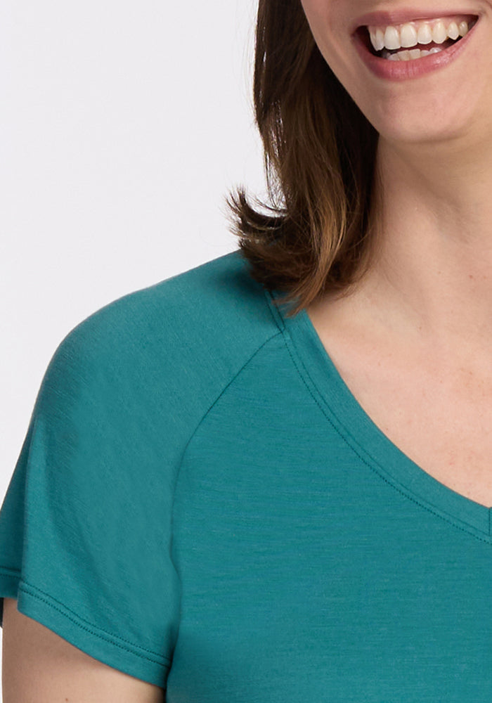 A close-up of a smiling woman wearing the Woolx Mia V Neck - Surfside tee. Only her mouth, part of her hair, neck, shoulder, and upper arm are visible against a plain white background.