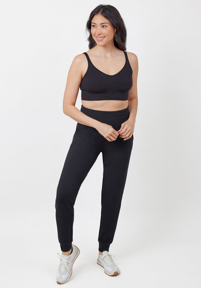 A woman with long dark hair stands against a white background, smiling to the side in Woolx Madden Joggers - Black, paired with a black sports bra and white sneakers.