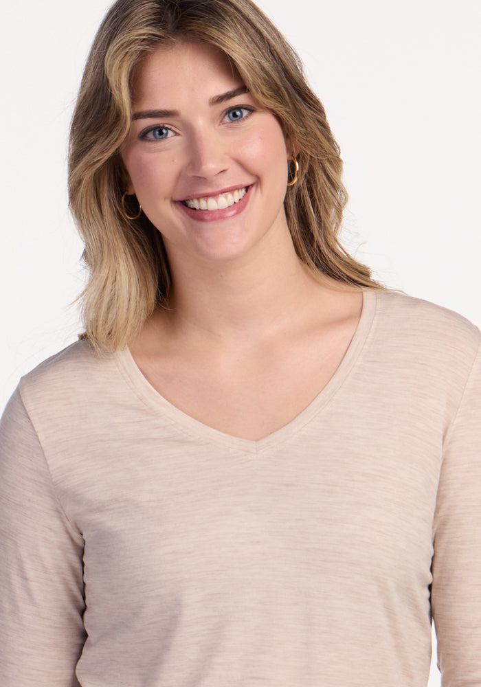 A young woman with shoulder-length blonde hair smiles at the camera against a plain white background, wearing the Woolx Millie Long Sleeve - Cream Heather and gold hoop earrings.