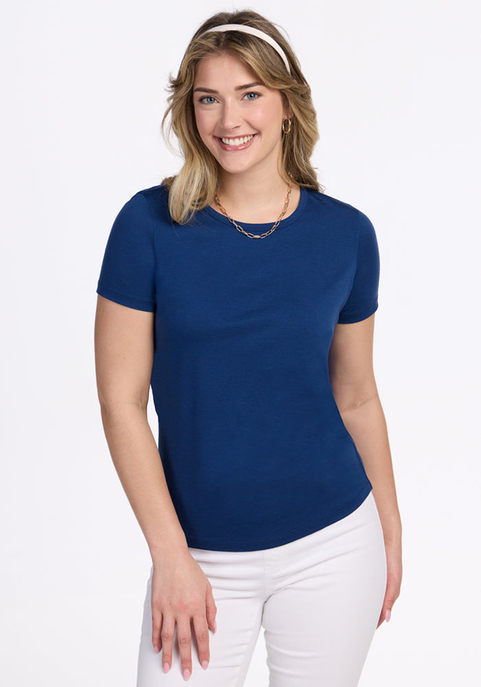 A woman with shoulder-length blonde hair wears the Woolx Liza Crew Neck Tee in Estate Blue, paired with white pants, a headband, and a gold necklace, and smiles at the camera against a plain white background.