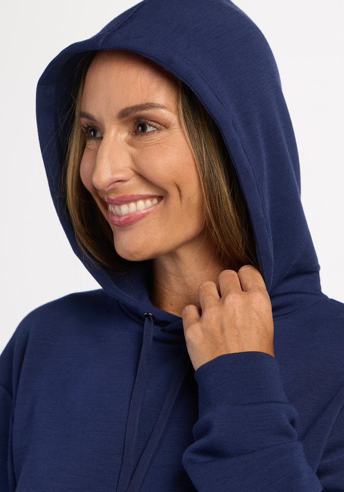 Smiling woman in a Woolx Avery Hoodie - Ocean Cavern, hood up, holding the cozy front edge with her right hand.