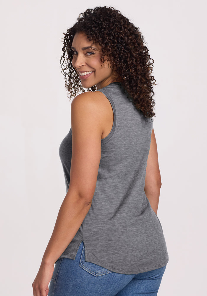 A woman with curly hair, wearing the Woolx Bella Tunic Top in Graphite Heather and blue jeans, smiles over her shoulder against a plain light background.