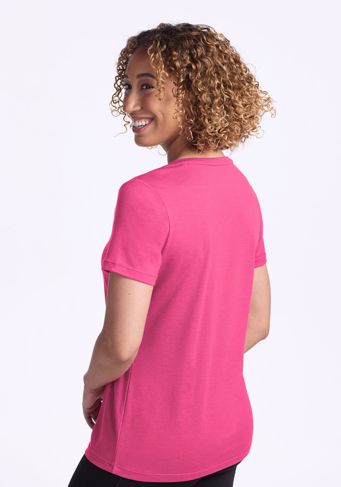 A person with curly hair wears the Woolx Junie V Neck in Raspberry Fizz, standing and smiling over their shoulder against a plain white background.