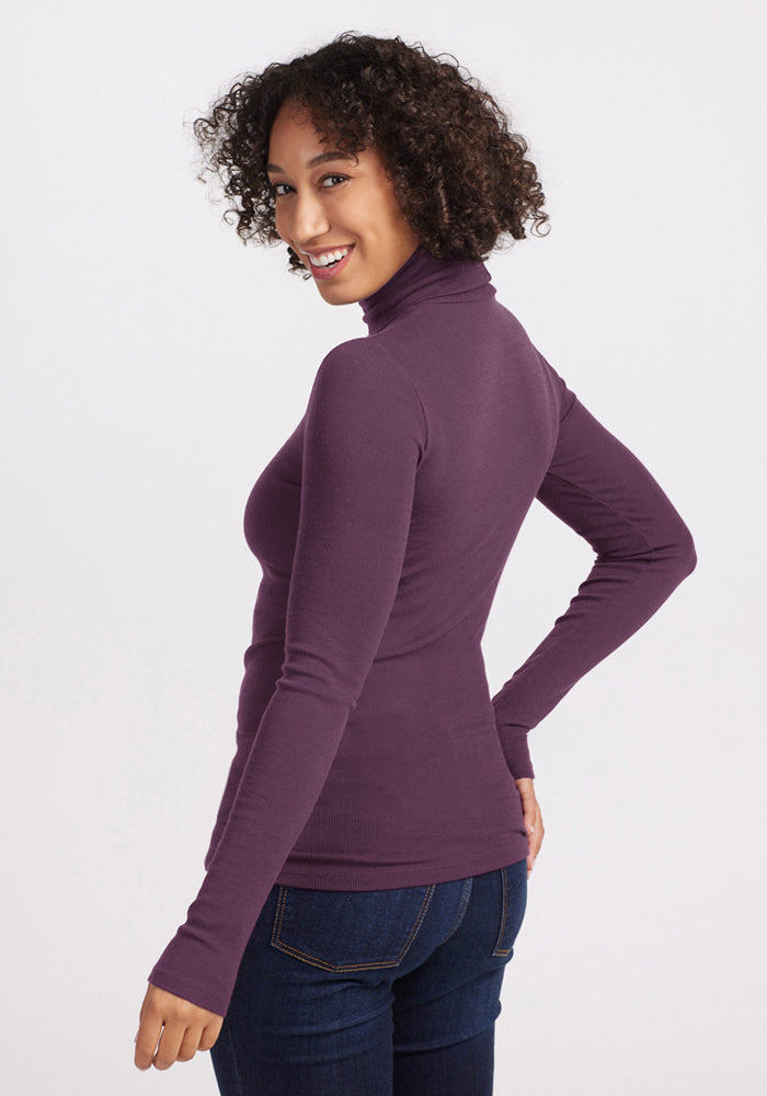 Woman with curly hair smiles in the Woolx Sage Ribbed Turtleneck - Purple Iris, posing with her back slightly turned.