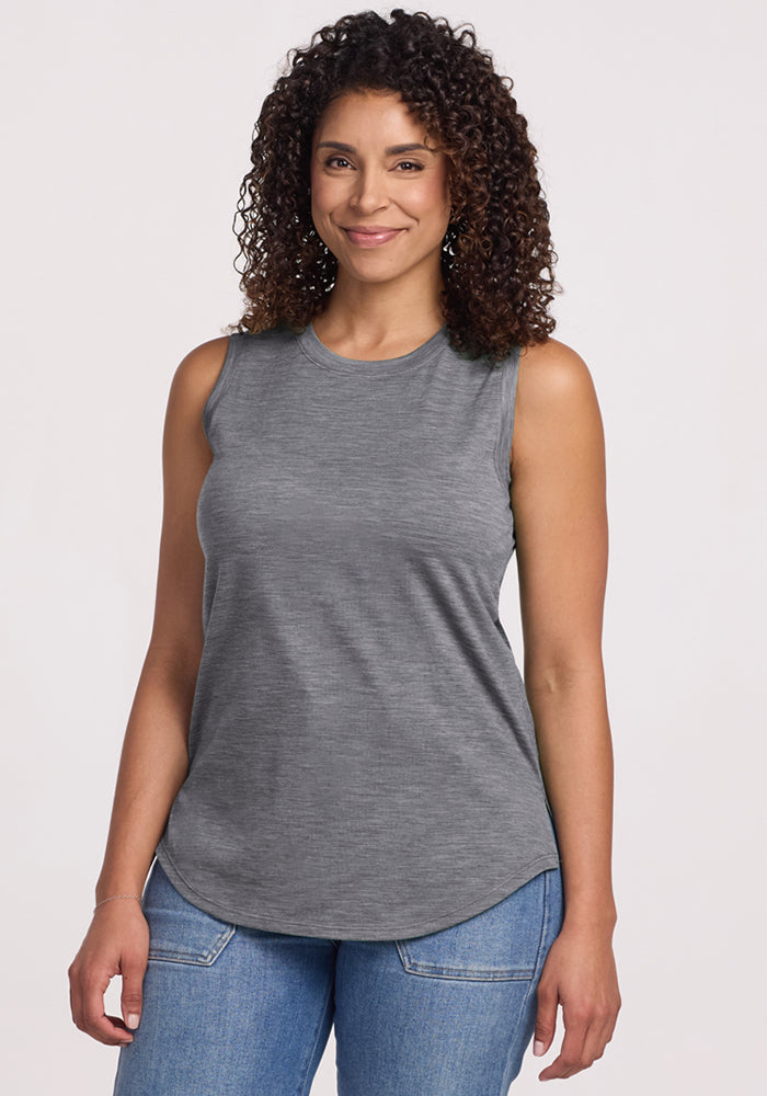 A woman with curly hair, smiling, wears the Woolx Bella Tunic Top in Graphite Heather and blue jeans against a plain light background. 