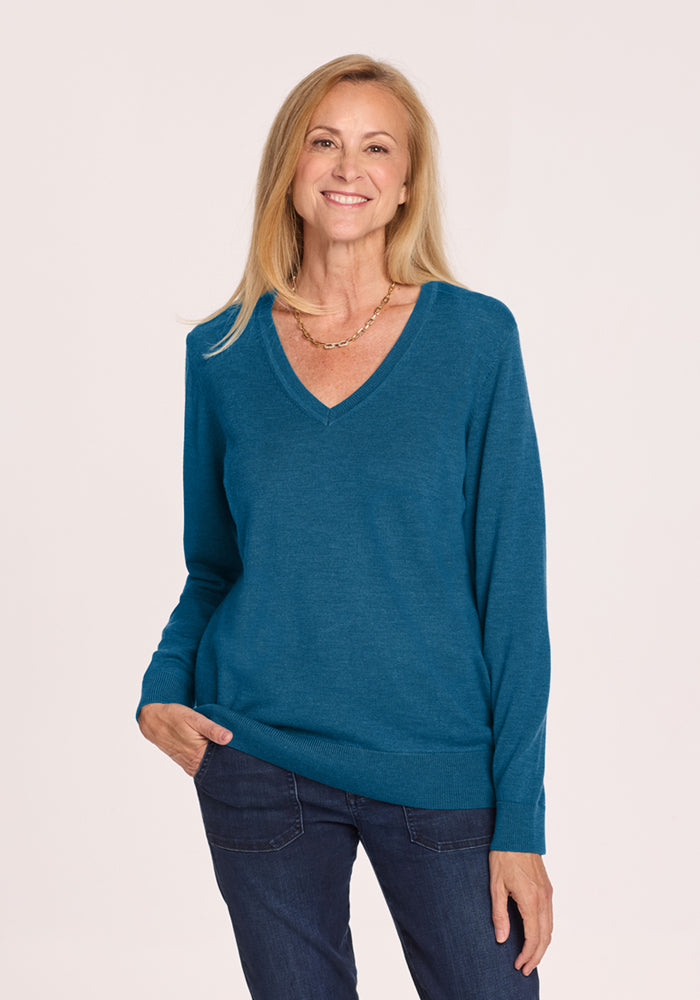 Smiling woman in the Woolx Francesca Sweater - Blue Pine and jeans stands against a plain background. 