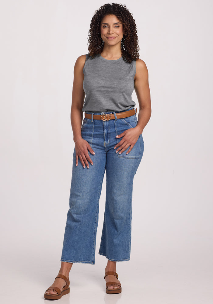 A woman with curly hair smiles, wearing the Woolx Bella Tunic Top in Graphite Heather, blue high-waisted jeans with a brown belt, and brown sandals, posing against a plain light background.