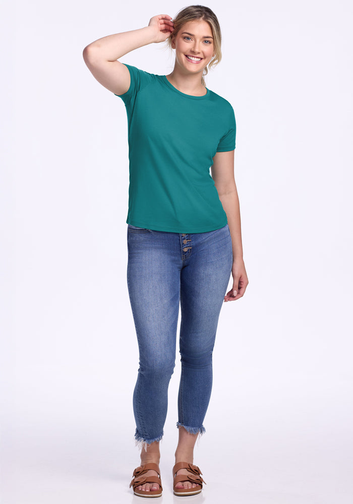 A smiling woman stands against a plain background, wearing the Woolx Liza Crew Neck Tee in Surfside, blue frayed-hem skinny jeans, and brown sandals. One hand touches her hair.