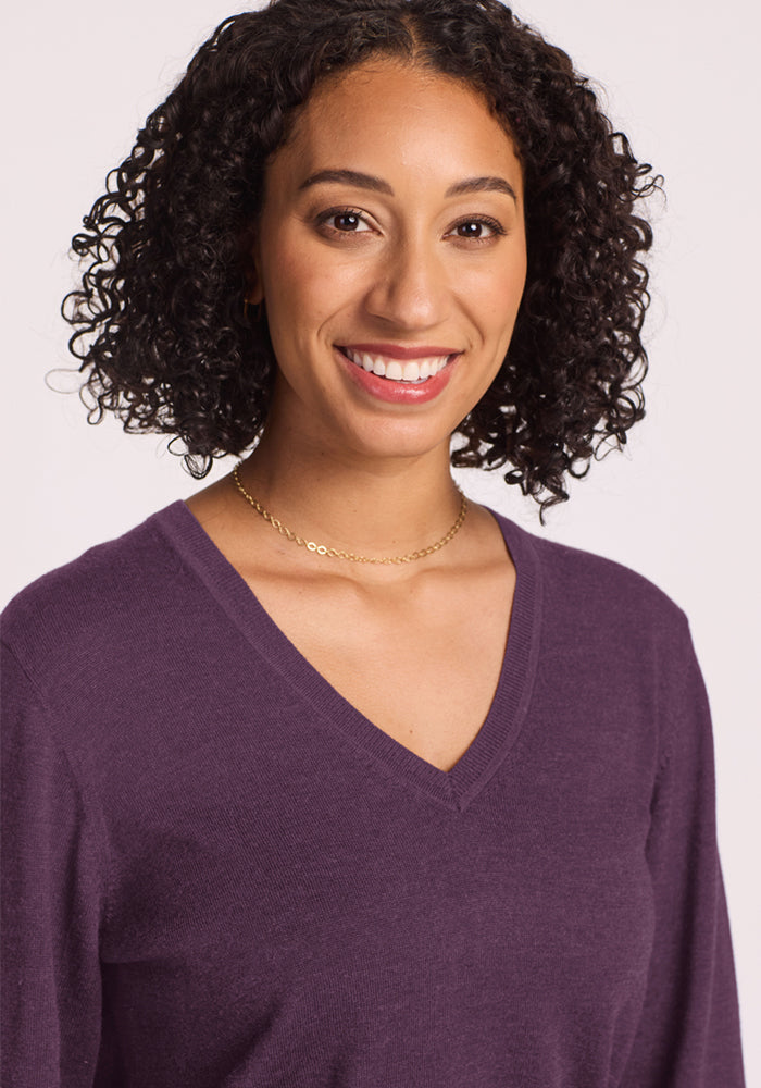 Smiling woman with curly hair wears a Woolx Francesca Sweater in Purple Iris and a gold necklace against a light background.