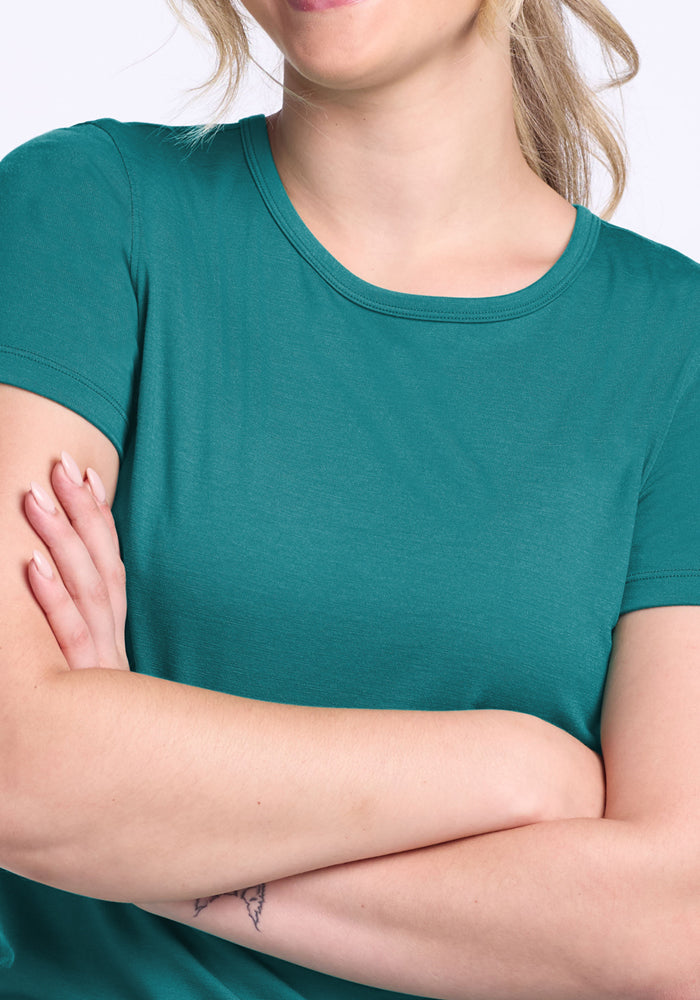 A blonde woman smiles with her arms crossed, showing a forearm tattoo, and wears the Woolx Liza Crew Neck Tee in Surfside. Her face is partially out of frame.