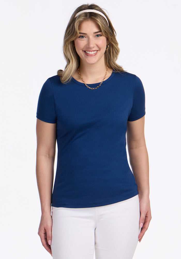 A smiling woman with shoulder-length blonde hair wears the Woolx Liza Crew Neck Tee in Estate Blue, a gold necklace, and white pants, standing against a plain white background. 